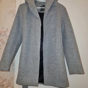 Grå kappa med luva  - Snygg grå kappa med luva och knytbälte i midjan. Perfekt för kyliga dagar med sin stilrena design och långa ärmar. Ett måste för garderoben!