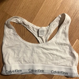 Vit sporttopp från Calvin Klein - Säljer en vit sporttopp från Calvin Klein med brottarrygg och elastiskt band med logotyp. Perfekt för träning eller som en bekväm vardagstopp. Funkar som S, personligen har 75A 