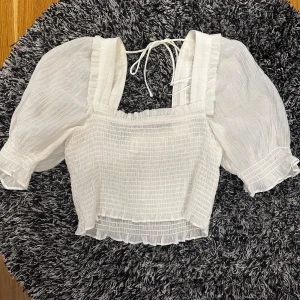 Vit blus med puffärmar - Charmig vit blus med puffiga kortärmar och knytband i ryggen. Den har en smockad design och volangdetaljer vid ärmslut och nederkant. Ändast använd en gång, så inga defekter🫶🏼 Den passar både S och M. Perfekt för sommarn!! 