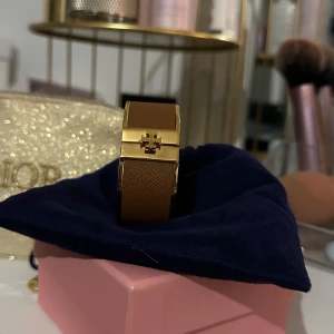 Brunt armband med gulddetaljer från Tory Burch - Elegant brunt armband med gulddetaljer från Tory Burch.Armbandet har en stilren design med ett guldspänne prytt med en logotyp. Perfekt för att ge en touch av lyx till din outfit.
