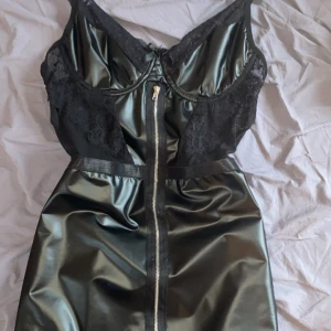 Svart klänning med spetsdetaljer - Snygg svart klänning med spetsdetaljer och dragkedja framtill. Klänningen har en glansig finish och är perfekt för en kväll ute. Den har en tight passform som framhäver figuren. Passar både xs/s