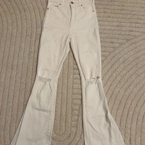 Vita bootcut jeans från Dr. Denim - Snygga vita bootcut jeans från Dr. Denim med hög midja och slitna detaljer vid knäna. Perfekta för en trendig look. De har en klassisk femficksdesign och knappgylf.
