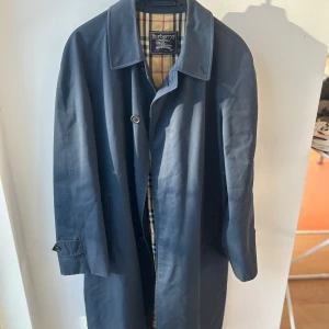 Blå trenchcoat från Burberry - Säljer en klassisk blå trenchcoat från Burberry med det ikoniska rutiga fodret. Jackan har långa ärmar och knappar framtill. Perfekt för en stilren look. Vintage, nyare verision säljs för ca 20 000kr.