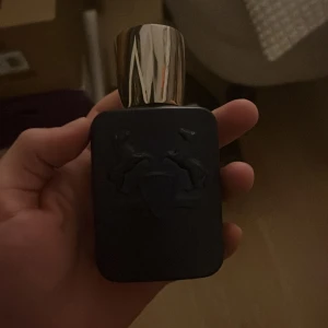 Layton Parfym från Parfums de Marly - Säljer en elegant Layton parfym från Parfums de Marly. Flaskan är mörkblå med en stilren design och har ett silverfärgat lock. Förpackningen är också mörkblå med vita detaljer och logotypen med två hästar. Perfekt för den som söker en lyxig doftupplevelse. 