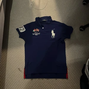 Blå pikétröja från Ralph Lauren - Säljer en blå( lite lila aktig) pikétröja från Ralph Lauren med stora vita och röda detaljer. Tröjan har korta ärmar och en klassisk krage. Perfekt för en sportig look med nummerdetaljer och texten 'ITALY' på baksidan.