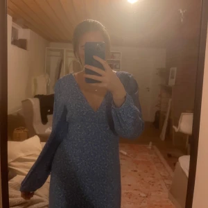 Blå blommig klänning - Säljer en söt blå klänning med blommigt mönster. Klänningen har en v-ringad design och långa ärmar, perfekt för vår och sommar. Den är i en normal passform och har en elegant längd. Köpt för 800kr om jag minns rätt, säljer för 300💗