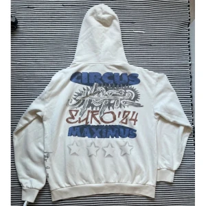 Travis Scott Hoddie  - Säljer en vit hoodie med unikt tryck på både fram- och baksida. Baksidan har texten 'CIRCUS EURO '84 MAXIMUS' i blått och rött med grafiska element. Framsidan har en stor illustration av en örn och textdetaljer. Perfekt för en avslappnad stil.