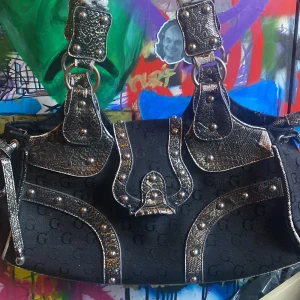 Svart axelväska med detaljer - Snygg svart axelväska med mönstrad yta och metalliska detaljer. Väskan har ett unikt mönster med bokstäver och är dekorerad med nitar och spännen i silverfärg. Typ y2k ju? 😍