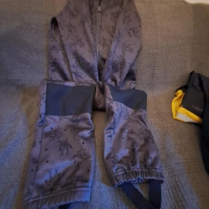 Grå barnbyxor med mönster från Lupilu - Grå barnbyxor från Lupilu med ett lekfullt mönster av olika figurer. Byxorna har dragkedja framtill och förstärkta knäpartier för extra hållbarhet. Perfekta för utomhuslek med justerbara fotremmar för bättre passform.