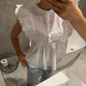 Vit blus med spetsdetaljer - Charmig vit blus med spetsdetaljer och volanger på ärmarna. Blusen har en knappslå framtill och ett broderat mönster som ger en romantisk känsla. Perfekt för en somrig look.
