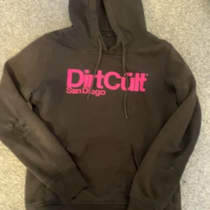Svart hoodie från DirtCult - Säljer en svart hoodie från DirtCult med rosa tryck 'San Diego' på framsidan. Tröjan har en klassisk design med huva och dragsko. Perfekt för en avslappnad stil. Den har dock hål vid armbågen på ena tröjarmen.