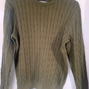 Stickad tröja från Tommy Hilfiger - Säljer en snygg olivgrön stickad tröja från Tommy Hilfiger i ekologisk bomull. Tröjan har ett klassiskt flätmönster och långa ärmar. Perfekt för kyliga dagar!