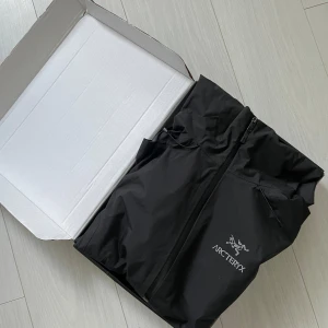 Svart regnjacka från Arc'teryx - Säljer en stilren svart regnjacka från Arc'teryx med dragkedja och huva. Jackan har broderad logotyp på bröstet och är perfekt för regniga dagar. Den är tillverkad i ett slitstarkt syntetmaterial.