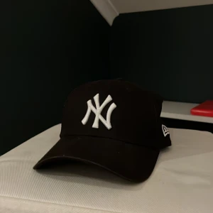 Svart keps från New Era - Snygg svart keps från New Era med den ikoniska NY-loggan broderad i vitt framtill. Kepsen är justerbar och tillverkad i 100% bomull, vilket gör den bekväm att bära. Perfekt för en casual look.