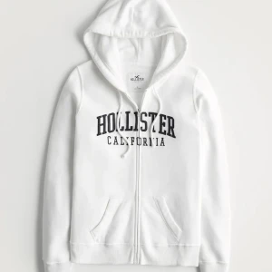 Vit Hollister hoodie - Säljer en vit hoodie från Hollister med dragkedja och texten 'Hollister California' i svart på framsidan. Den har en justerbar huva och två fickor framtill. Bra skick men kommer inte till användning. Skriv om du har frågor eller vill ha fler bilder!💘