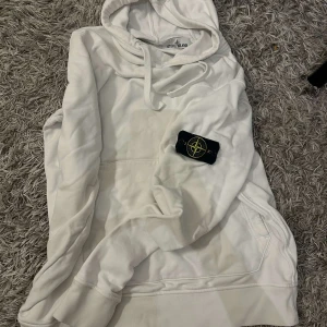 Vit hoodie från Stone Island - Säljer en stilren vit hoodie från Stone Island med den ikoniska loggan på ärmen. Hoodien har en klassisk design med dragsko i huvan och en stor ficka framtill. Perfekt för en avslappnad stil.