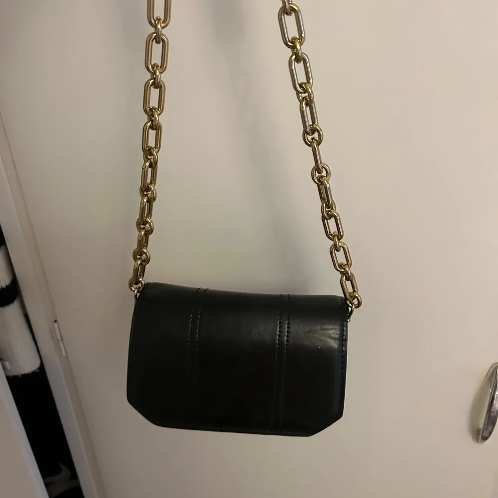 Snygg svart axelväska i skinn med guldfärgad kedja som axelrem. Väskan har en praktisk dragkedja och en knappstängning framtill. Aldrig använd, så tänker kan komma till nytta hos någon annan!☺️. Laukut & Käsilaukut.
