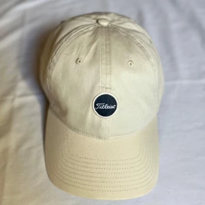 Beige keps från Titleist - Snygg beige keps från Titleist med en broderad logotyp framtill. Perfekt för golfsäsongen. Justerbar passform för optimal komfort. Helt ny. Bara att höra av sig vid frågor//Carl