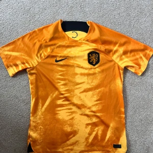 Orange fotbollströja från Nike - Snygg orange fotbollströja från Nike med korta ärmar och ett svart lejonemblem på bröstet. Perfekt för fotbollsfans som vill sticka ut på planen eller läktaren. Tröjan har en modern design med en skön passform.