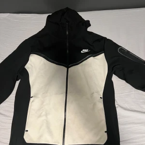 Svart och vit jacka från Nike - Säljer en snygg svart och vit jacka från Nike med dragkedja och huva. Jackan har en sportig design med långa ärmar och en liten Nike-logga på bröstet. Perfekt för en aktiv livsstil.