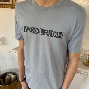 Ljusblå t-shirt från Woolrich - Säljer en stilren ljusblå t-shirt från Woolrich med svart tryck på bröstet. Perfekt för en avslappnad look. T-shirten har en klassisk passform och är kortärmad.