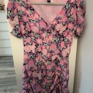 Blommig klänning från H&M  - Säljer en blommig klänning från H&M  i storlek 34. Klänningen har ett färgglatt mönster i rosa, lila och blå toner med korta ärmar och en v-ringad hals. Den kan spännas in/ut på längden och en har en dragkedja på sidan.