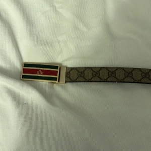 Mönstrat bälte från Gucci - Snyggt bälte från Gucci med klassiskt GG-mönster i beige och brunt. Spännet är rektangulärt med gröna och röda detaljer samt en guldig finish. Perfekt för att ge en lyxig touch till din outfit. De går däremot att ändra färg på den till svart och beige säljer den för att ja aldrig har använt den fick den av en släkting 