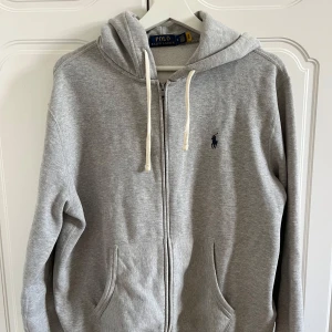 Grå hoodie från Polo Ralph Lauren - Säljer en stilren grå hoodie från Polo Ralph Lauren med dragkedja och justerbar huva. Tröjan har en broderad logga på bröstet och praktiska fickor framtill. Perfekt för en avslappnad look.