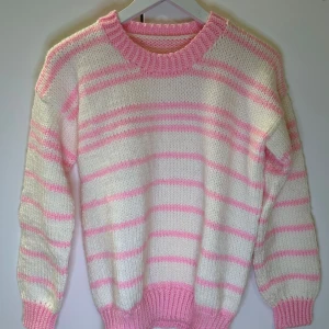 Rosa och vit randig stickad tröja - Mysig handstickad tröja i rosa och vitt med randigt mönster. Tröjan har långa ärmar och ribbade muddar vid ärmslut och nederkant. Perfekt för kyliga dagar!
