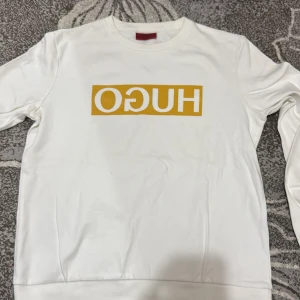 Vit tröja från Hugo - Säljer en stilren vit tröja från Hugo med ett gult tryck på framsidan. Tröjan har långa ärmar och en klassisk rund halsringning. Perfekt för en avslappnad look.