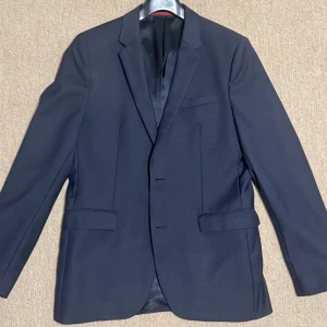  Hugo Boss kavaj  - Elegant svart kavaj från Hugo Boss i klassisk design. Perfekt för formella tillfällen med en stilren look. Kavajen har en enkelknäppt front och fickor med lock. Den är endast använd ett fåtal gånger och i bra skick, storlek 50 som motsvarar en M/L