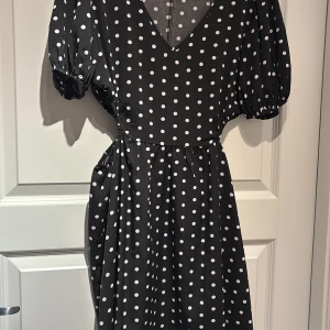 Svart prickig klänning med volangdetaljer - Trendig svart klänning med vita prickar och volangdetaljer. Den har en djup v-ringning och korta ärmar med volanger. Perfekt för en somrig look.