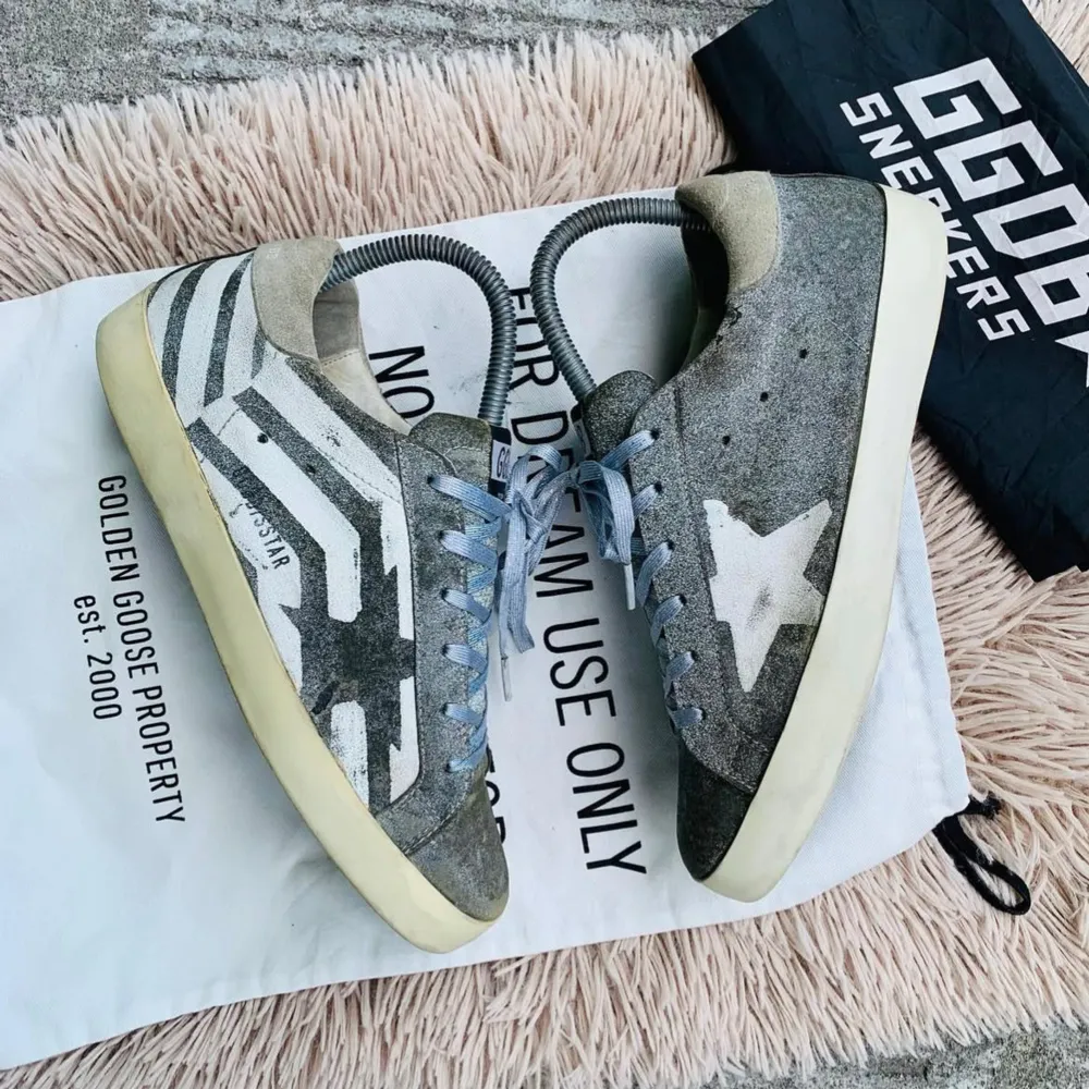 Snygga grå sneakers från Golden Goose med vit stjärndetalj och randigt mönster på sidan. Skorna har ljusblå snörning och en krämfärgad sula. Perfekta för en avslappnad stil med en touch av unik design.. Kengät.