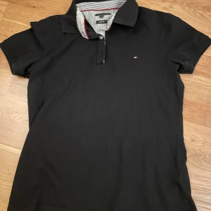 Svart kortärmad pikétröja från Tommy Hilfiger - Snygg svart pikétröja från Tommy Hilfiger i slim fit. Tröjan har korta ärmar och en klassisk krage med randigt mönster på insidan. Perfekt till sommaren! Storlek XXL för barn vilket är ~XS herr.