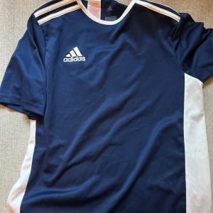 Mörkblå fotbollströja från Adidas - Snygg mörkblå fotbollströja från Adidas med vita detaljer och korta ärmar. Tröjan har de klassiska tre ränderna på axlarna och Adidas-loggan på bröstet. Perfekt för träning eller match!