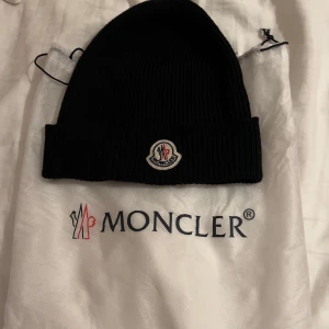Svart ribbad mössa från Moncler - Säljer en snygg svart ribbad mössa från Moncler. Den är i nyskick och perfekt för kalla dagar. Mössan har Monclers ikoniska logga framtill och är supermjuk och bekväm. Passar perfekt till både vardag och vinteräventyr! Mössan är äkta!