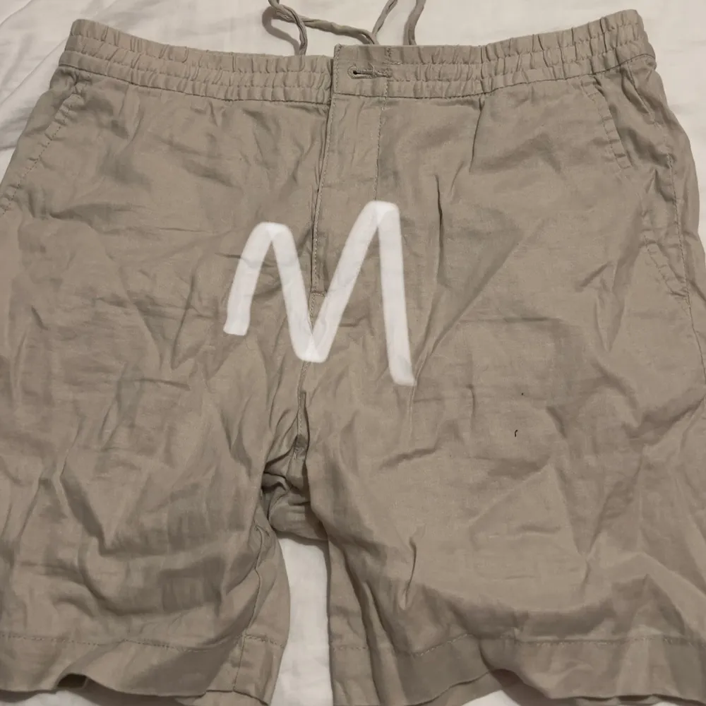 Säljer ett par bekväma beige shorts i linne. Perfekta för sommardagar med sin lätta och luftiga känsla. De har en elastisk midja med dragsko för justerbar passform. Storlek M, passar både för avslappnade dagar och strandhäng.. Shorts.