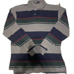 Randig pikétröja från Ralph Lauren - Säljer en klassisk pikétröja från Ralph Lauren i storlek XL. Den har ett randigt mönster i grått, blått och grönt med röda detaljer. Tröjan är långärmad och har en broderad logga på bröstet. Perfekt för en avslappnad men stilren look! Fråga gärna om extra bilder eller prisförslag 👍