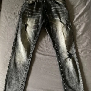 Grå jeans med slitningar - Säljer ett par grå jeans med coola slitningar på framsidan. De har en klassisk femficksdesign och en snygg tvättad look. Perfekta för en avslappnad stil och passar till det mesta. Sköna och trendiga för både vardag och fest!