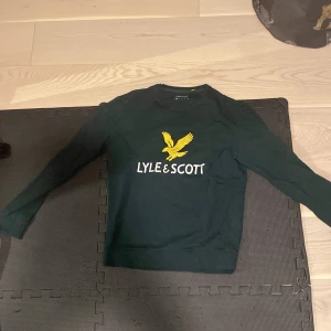 Mörkgrön tröja från Lyle & Scott - Säljer en snygg mörkgrön tröja från Lyle & Scott i storlek S. Tröjan är gjord av 100% bomull och har en klassisk design med det ikoniska gula örnlogotypen på bröstet. Perfekt för både vardag och lite finare tillfällen. Skön och stilren, passar till det mesta! 💚