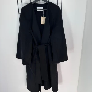 Stylein Kappa Trento Svart - Kimono inspirerad kappa med tappade axlar och utsvängda armar i full längd med en subtit sömstil framtill. Ett fäst bälte i midjan som skapar mångisidighet i utseende. Tillverkad av ullblandning.  Kappan är helt oanvänd med prislapp kvar. Storkel M  