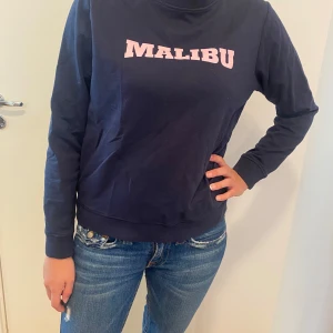 Mörkblå sweatshirt med Malibu-tryck - Säljer en snygg mörkblå sweatshirt med rosa 'Malibu'-tryck på bröstet. Tröjan är i bomull och har långa ärmar, perfekt för en avslappnad stil. Den är i bra skick och passar perfekt till jeans för en casual look. Perfekt för höst och vår!