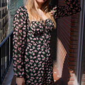 Blommig klänning från ADOORE - Säljer en superfin blommig klänning från ADOORE i storlek 34. Den har ett svart tyg med rosa blommor och långa ärmar. Klänningen har en söt knytning framtill och en liten slits som ger en extra touch. Perfekt för vår och sommar! 🌸