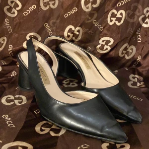 Svarta klackskor från Zara - Snygga svarta klackskor från Zara i skinn med spetsig tå och öppen häl. De har en elegant klack med en unik detalj runtom. Perfekta för både fest och vardag. Skorna är i bra skick och redo för nya äventyr!