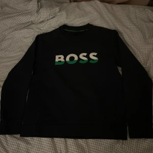 Svart tröja från Boss - Säljer en stilren svart tröja från Boss med deras logga i vitt och grönt på framsidan. Tröjan har långa ärmar och är perfekt för en casual look. Passar bra till både jeans och chinos. Perfekt för höst och vinter!