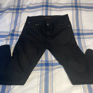 Svarta Nudie Jeans W31 L34 - Säljer ett par svarta jeans från Nudie Jeans med diskret broderi på bakfickorna. Jeansen har en slim passform och är tillverkade i mjukt bomullstyg med lite stretch för extra komfort. Perfekta för dig som gillar stilrena och enkla jeans. Riktigt schtek!