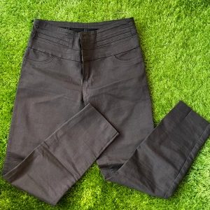 Bruna högmidjade byxor med knappar - Snygga mörkgrå byxor med hög midja och flera dekorativa sömmar upptill. Byxorna har fyra synliga knappar och dragkedja framtill, samt raka ben och diskreta bakfickor. Perfekta för en clean och stilren look.