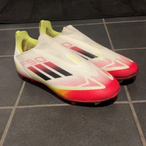 Adidas F50 vita och röda fotbollsskor - Säljer ett par Adidas F50 fotbollsskor i vitt med röda och gula detaljer samt svarta ränder. Skorna har en strömlinjeformad design utan snörning och är tillverkade i syntetmaterial. Sulan är röd med gula dobbar för bra grepp på planen.