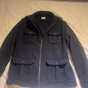 Blå cashmere/ulljacka från John Henric - Stilren blå jacka från John Henric med fyra stora fickor framtill och dragkedja. Jackan är tillverkad i en lyxig mix av ull och kashmir, vilket gör den både mjuk och varm. Perfekt för dig som gillar klassisk och clean look.