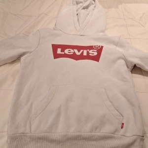 Vit hoodie från Levi's med röd logga - Vit hoodie från Levi's med klassisk röd logga framtill. Tröjan har huva, magficka och ribbade muddar vid ärmslut och nederkant. Perfekt för dig som gillar en clean och stilren look med streetkänsla.